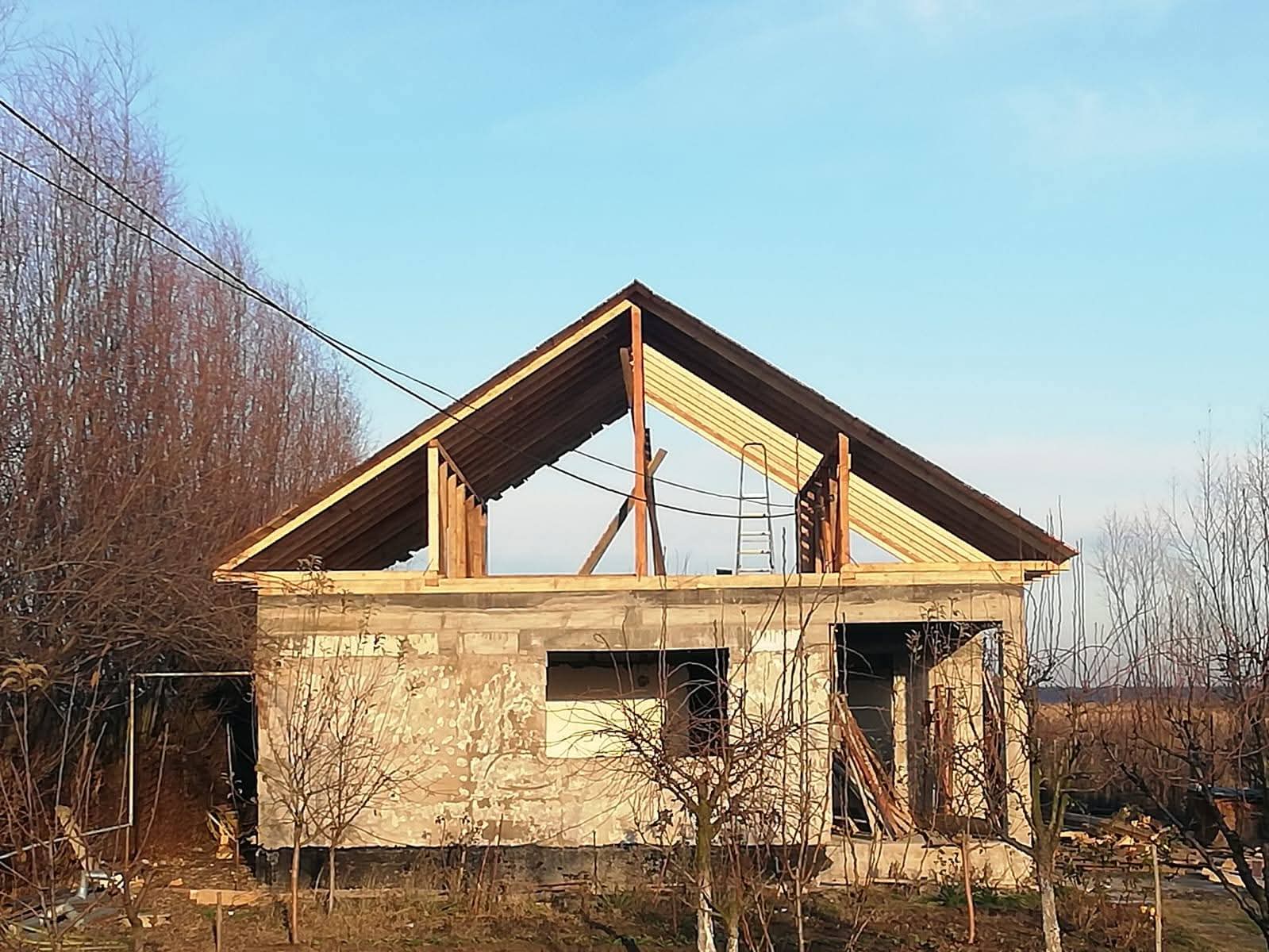 Charpente et gros œuvre : Avancement de la construction d’une maison. Maison en construction : gros œuvre et charpente de toit en pignon installés. Murs bruts et ciel bleu sur fond d'arbres dénudés.