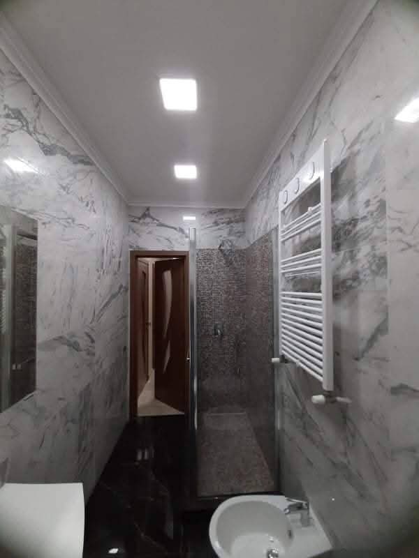 Salle de bain moderne en marbre blanc et noir avec douche Salle de bain moderne étroite en marbre blanc et noir. Douche à l'italienne, bidet et radiateur sèche-serviettes. Lumières carrées au plafond.