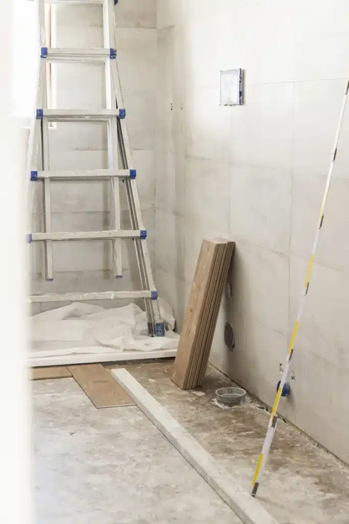 Chantier de rénovation : pose de carrelage et sol en attente. Rénovation intérieure : murs carrelés clairs, échelle, sol en béton avec lattes de bois pour parquet et outils de mesure.